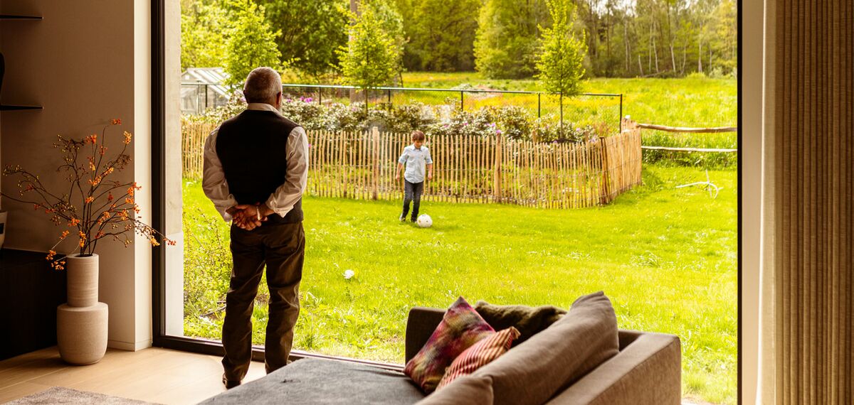 Elder person watching his grandson play in the garden from behind window wall.