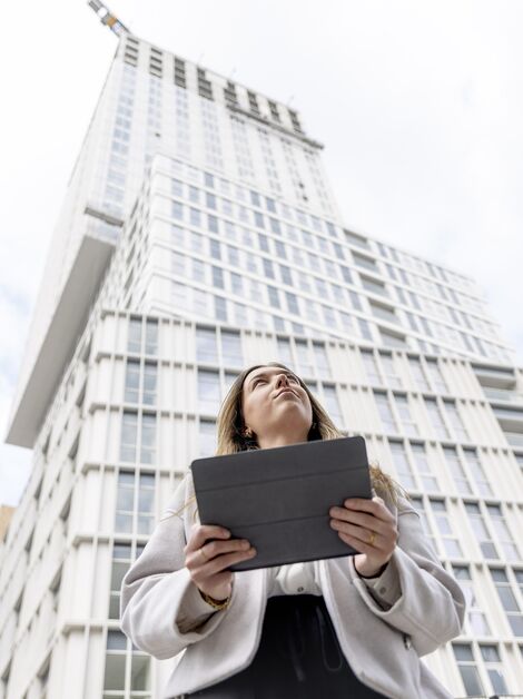 Blonďatá žena s iPadem v ruce se dívá směrem k bílému obytnému domu.